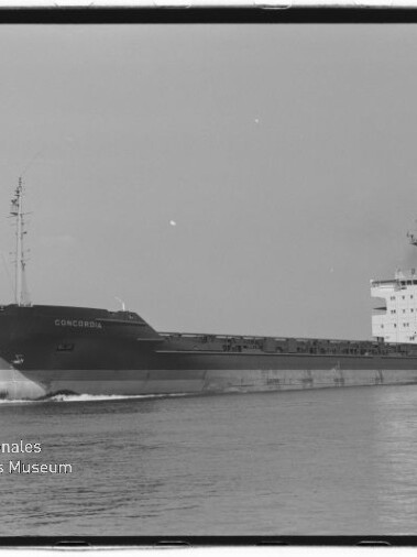 Schwarzweißaufnahme eines Frachtschiffs mit dem Namen CONCORDIA, seitliche Ansicht beim Fahren auf ruhigem Wasser mit langem Laderaum und Aufbauten achtern; Ufer mit Bäumen im Hintergrund und Museumswasserzeichen unten links.
