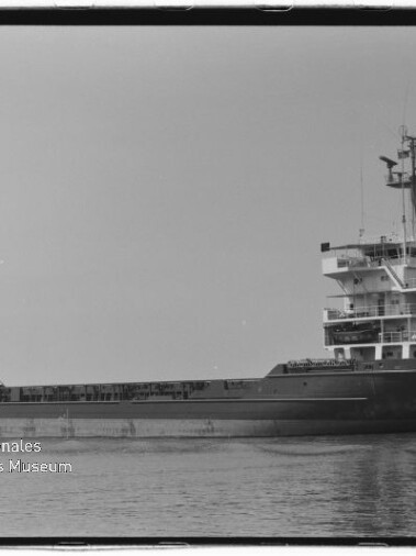 Schwarzweiß-Seitenaufnahme eines Frachtschiffs mit hohem Aufbau achtern, offener Ladefläche und kurzem Mast vorn, das ruhig auf glattem Wasser fährt; unten links ist das Logo des Internationalen Maritimen Museums Hamburg sichtbar.