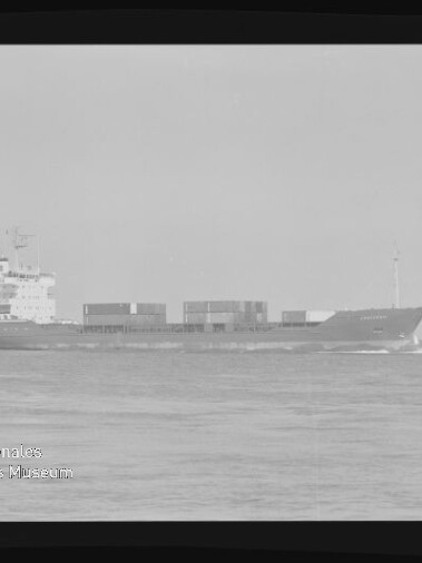 Schwarzweiß-Aufnahme eines Containerschiffs in Seitenansicht auf ruhiger See mit gestapelten Containern auf dem Deck, dem Aufbauten/Brückenturm hinten links, sichtbaren Filmrändern und einem Museumswasserzeichen unten links.