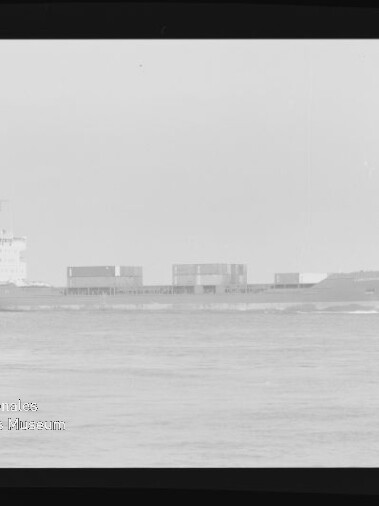 Schwarzweiß-Foto eines Containerschiffs in Seitenansicht (Bug nach rechts) auf ruhiger See, mit mehreren Containern an Deck und Aufbauten am Heck; schwarzer Filmrand und Museumslogo unten links sichtbar.