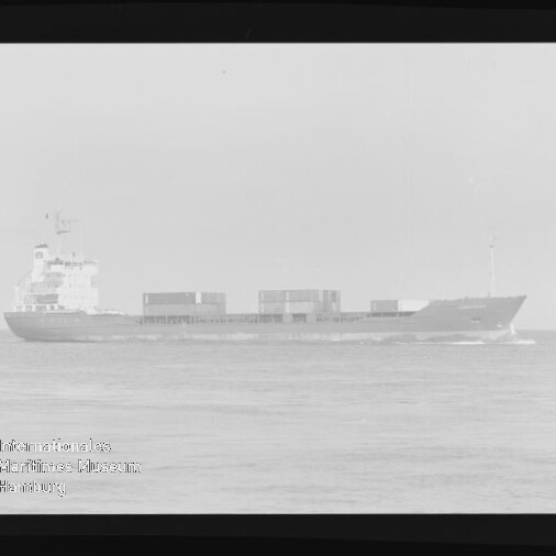 Schwarzweiß-Foto eines Containerschiffs in Seitenansicht (Bug nach rechts) auf ruhiger See, mit mehreren Containern an Deck und Aufbauten am Heck; schwarzer Filmrand und Museumslogo unten links sichtbar.