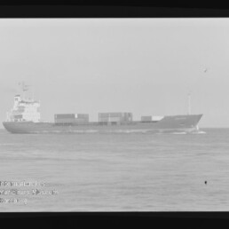 Schwarzweiß-Fotografie eines Containerschiffs in Seitenansicht auf ruhiger See mit mehreren auf dem Deck gestapelten Containern, leichter Bugwelle, sichtbarem Filmrand und Wasserzeichen des Internationalen Maritimen Museums Hamburg.