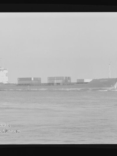 Schwarzweiß-Fotografie eines Containerschiffs in Seitenansicht auf ruhiger See mit mehreren auf dem Deck gestapelten Containern, leichter Bugwelle, sichtbarem Filmrand und Wasserzeichen des Internationalen Maritimen Museums Hamburg.