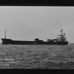 Schwarzweiß-Seitenansicht eines Frachtschiffs mit der Aufschrift CJS auf dem Rumpf und Containern am Heck, das auf leicht gewellter See fährt; links unten Wasserzeichen des Internationalen Maritimen Museums Hamburg.