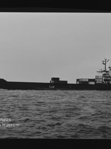Schwarzweiß-Seitenansicht eines Frachtschiffs mit der Aufschrift CJS auf dem Rumpf und Containern am Heck, das auf leicht gewellter See fährt; links unten Wasserzeichen des Internationalen Maritimen Museums Hamburg.