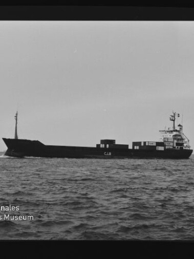 Schwarzweiß-Foto eines Frachtschiffs in Seitenansicht auf ruhiger See mit wenigen Containern an Deck; sichtbarer Filmrand und -nummern sowie links unten das Wasserzeichen des Internationalen Maritimen Museums Hamburg.