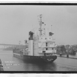 Schwarzweißaufnahme eines Frachtschiffs mit der Beschriftung Alice Bülten, das in einem Hafenbecken an Kais und Hafenkränen vorbeifährt; im Hintergrund eine Brücke und unten links das Wasserzeichen des Internationalen Maritimen Museums Hamburg.