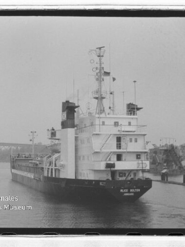 Schwarzweißaufnahme eines Frachtschiffs mit der Beschriftung Alice Bülten, das in einem Hafenbecken an Kais und Hafenkränen vorbeifährt; im Hintergrund eine Brücke und unten links das Wasserzeichen des Internationalen Maritimen Museums Hamburg.