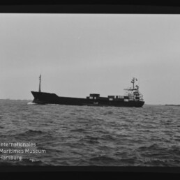 Schwarzweißaufnahme eines Frachtschiffs in Seitenansicht auf leicht bewegter See, mit aufragender Brücke achtern, Containern auf dem Deck, sichtbarem Filmrand ‚AGFAPAN 100‘ und Wasserzeichen des Internationalen Maritimen Museums Hamburg.