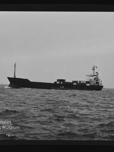 Schwarzweißaufnahme eines Frachtschiffs in Seitenansicht auf leicht bewegter See, mit aufragender Brücke achtern, Containern auf dem Deck, sichtbarem Filmrand ‚AGFAPAN 100‘ und Wasserzeichen des Internationalen Maritimen Museums Hamburg.