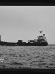 Schwarzweiß-Foto eines Frachtschiffs in Seitenansicht auf leicht welliger See mit wenigen Containern auf dem Deck und achtern liegendem Aufbauten, sichtbarem Filmrand mit der Markierung „12“ links sowie dem Logo des Internationalen Maritimen Museums Hamburg in der linken unteren Ecke.