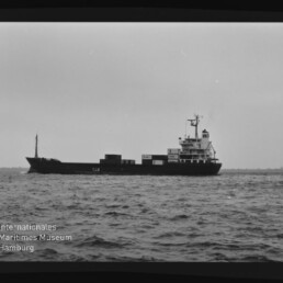 Schwarzweiß-Foto eines Frachtschiffs in Seitenansicht auf leicht welliger See mit wenigen Containern auf dem Deck und achtern liegendem Aufbauten, sichtbarem Filmrand mit der Markierung „12“ links sowie dem Logo des Internationalen Maritimen Museums Hamburg in der linken unteren Ecke.