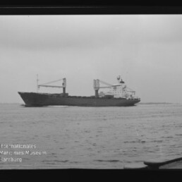 Schwarzweißaufnahme eines seitlich gezeigten Frachtschiffs mit zwei auf Deck stehenden Ladekränen, das bei ruhiger See am Horizont vorbeifährt; links unten das Logo und der Schriftzug des Internationalen Maritimen Museums Hamburg.
