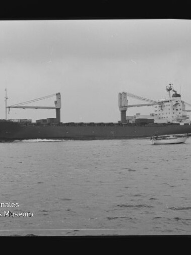Schwarzweiß-Foto eines Frachtschiffs mit zwei Ladekränen, das auf ruhiger See fährt; im Vordergrund ein kleines Motorboot, unten links Wasserzeichen des Internationalen Maritimen Museums Hamburg und sichtbarer Filmrand.