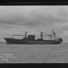 Schwarzweiß-Foto eines Containerschiffs mit der Aufschrift CMB, beladen mit Containern und zwei Ladekränen, das auf ruhiger See unter bedecktem Himmel fährt; unten links Wasserzeichen des Internationalen Maritimen Museums Hamburg.