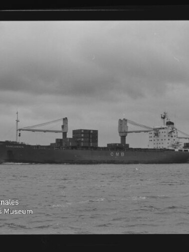 Schwarzweiß-Foto eines Containerschiffs mit der Aufschrift CMB, beladen mit Containern und zwei Ladekränen, das auf ruhiger See unter bedecktem Himmel fährt; unten links Wasserzeichen des Internationalen Maritimen Museums Hamburg.