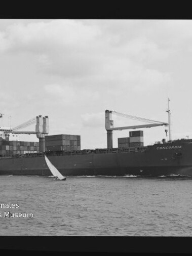 Schwarzweißaufnahme eines Containerschiffs namens „Concordia“ mit gestapelten Containerboxen und zwei Ladekränen, das auf dem Meer fährt und von kleinen Segelbooten begleitet wird; Filmrand mit Aufschrift AGFA APX 100 und Museumswasserzeichen unten links.