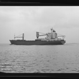 Schwarzweiß-Seitenansicht eines Frachtschiffs mit aufgesetzten Ladekränen auf ruhiger See und bedecktem Himmel, unten links das Wasserzeichen des Internationalen Maritimen Museums Hamburg.