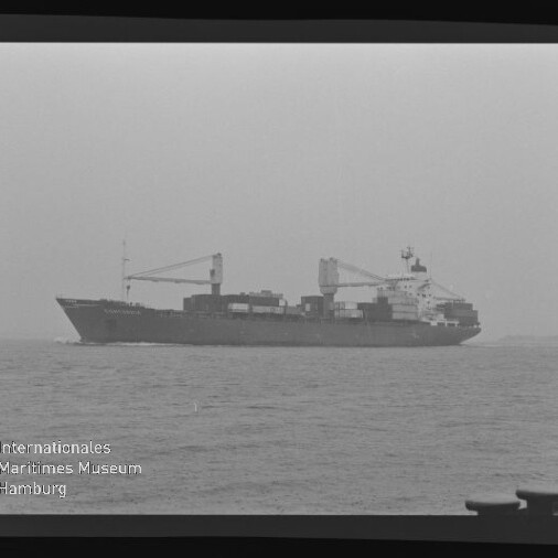 Schwarz-weiß-Foto eines Containerfrachters mit zwei Ladekränen und gestapelten Containern auf ruhiger, leicht nebliger See; sichtbarer Filmrand und Wasserzeichen des Internationalen Maritimen Museums Hamburg in der linken unteren Ecke.