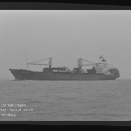 Seitliche Schwarzweißaufnahme eines Containerschiffs mit Ladekränen und gestapelten Containern auf ruhiger See unter grauem Himmel, mit Wasserzeichen Internationales Maritimes Museum Hamburg unten links.