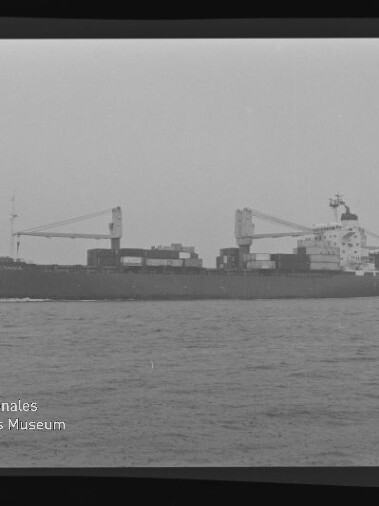 Schwarzweiß-Seitenansicht eines Containerschiffs mit aufgestapelten Containern und zwei Ladekränen, das bei ruhiger See von links nach rechts fährt; unten links Logo und Schriftzug des Internationalen Maritimen Museums Hamburg.