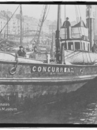 Schwarzweißaufnahme eines am Kai vertäuten Schiffes mit dem Schriftzug CONCURRENT am Bug, mehreren Personen an Bord und einer Hafenkulisse mit Bootsmasten und Häusern im Hintergrund; links unten Wasserzeichen des Internationalen Maritimen Museums Hamburg.