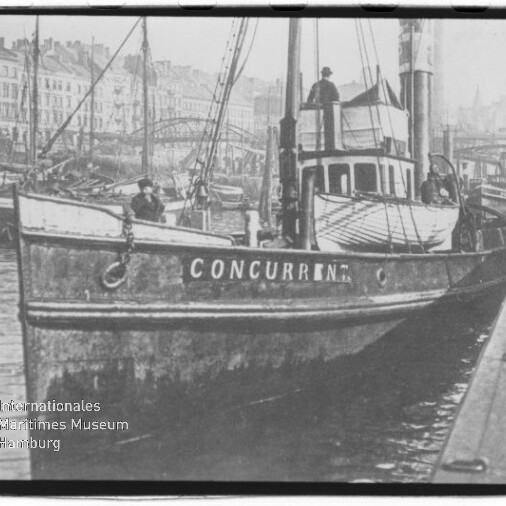 Schwarzweißaufnahme eines am Kai vertäuten Schiffes mit dem Schriftzug CONCURRENT am Bug, mehreren Personen an Bord und einer Hafenkulisse mit Bootsmasten und Häusern im Hintergrund; links unten Wasserzeichen des Internationalen Maritimen Museums Hamburg.