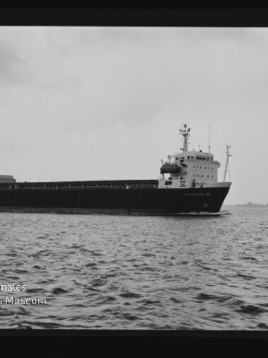Schwarzweiß-Aufnahme eines Frachtschiffs in Seitenansicht auf ruhiger See vor bewölktem Himmel, im Vordergrund welliges Wasser und unten links ein Logo mit der Aufschrift „Internationales Maritimes Museum Hamburg“.