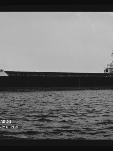 Schwarzweiß-Seitenansicht eines großen Massengutfrachters auf ruhiger See mit langem Ladebereich und achtern gelegenem Steuerhaus, unten links Wasserzeichen des Internationalen Maritimen Museums Hamburg.