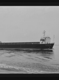 Schwarz-weiß-Aufnahme eines langen Frachtschiffs, das ruhig über eine spiegelnde Wasseroberfläche fährt; Steuerhaus und Aufbauten vorne rechts sichtbar, flacher Horizont im Hintergrund und Museumswasserzeichen unten links.