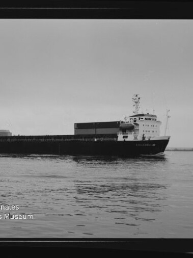 Schwarzweiß-Foto eines Frachtschiffs in Seitenansicht auf ruhigem Wasser, mit sichtbarem Aufbau, geladenen Deckseinheiten, weiter Horizont und dem Logo des Internationalen Maritimen Museums Hamburg unten links.