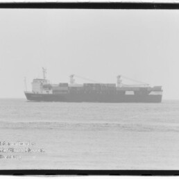 Schwarzweißaufnahme eines Containerschiffs im Profil auf ruhiger See, mit gestapelten Containern und zwei Ladekränen, unscharfer Horizont und Wasserzeichen des Internationalen Maritimen Museums Hamburg unten links.