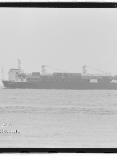 Schwarzweißaufnahme eines Containerschiffs im Profil auf ruhiger See, mit gestapelten Containern und zwei Ladekränen, unscharfer Horizont und Wasserzeichen des Internationalen Maritimen Museums Hamburg unten links.