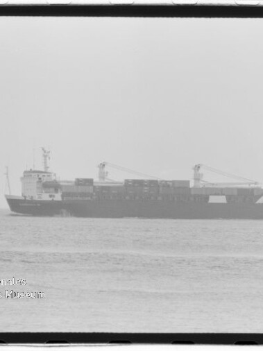 Schwarzweiß-Aufnahme eines Containerschiffs in Seitenansicht auf ruhigem Meer mit gestapelten Containern und zwei Ladekranen, unten links Wasserzeichen „Internationales Maritimes Museum Hamburg“ und sichtbarer Filmrand.