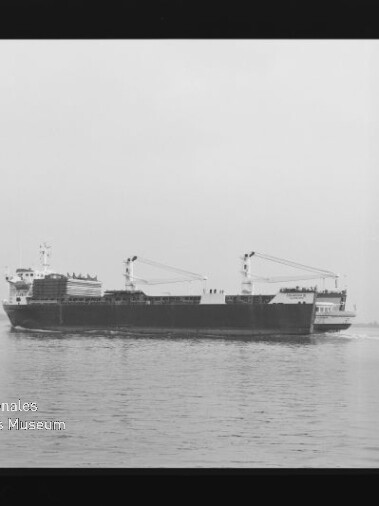 Schwarzweiß-Foto eines Frachtschiffs mit zwei Deckskränen und achterem Aufbauten, das ruhig auf dem Meer nahe der flachen Küstenlinie fährt; links unten sichtbar das Wasserzeichen des Internationalen Maritimen Museums Hamburg.
