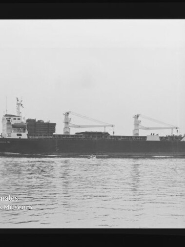 Schwarz-weiß-Aufnahme eines Frachtschiffs in Seitenansicht auf ruhiger See, mit zwei Deckkränen, gestapelter Ladung an Deck und sichtbarem Negativrand sowie Wasserzeichen des Internationalen Maritimen Museums Hamburg