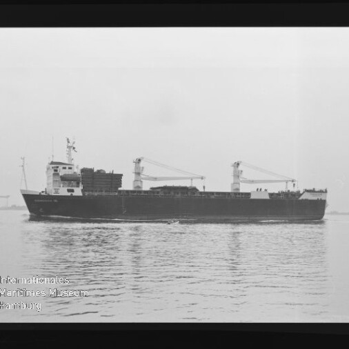 Schwarz-weiß-Aufnahme eines Frachtschiffs in Seitenansicht auf ruhiger See, mit zwei Deckkränen, gestapelter Ladung an Deck und sichtbarem Negativrand sowie Wasserzeichen des Internationalen Maritimen Museums Hamburg