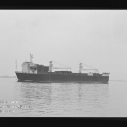 Schwarzweißaufnahme eines Frachtschiffs mit zwei Ladekränen, seitlich auf ruhigem Wasser fahrend; das Bild zeigt Filmrand und unten links ein Wasserzeichen des Internationalen Maritimen Museums Hamburg.