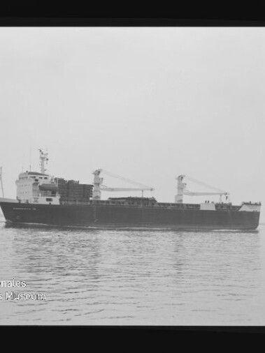 Schwarzweißaufnahme eines Frachtschiffs mit zwei Ladekränen, seitlich auf ruhigem Wasser fahrend; das Bild zeigt Filmrand und unten links ein Wasserzeichen des Internationalen Maritimen Museums Hamburg.