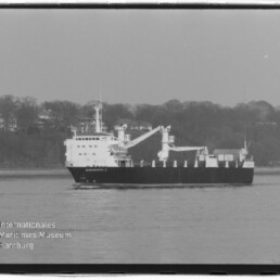 Schwarz-weiß-Foto eines Frachtschiffs mit zwei Ladekränen, das auf ruhigem Wasser vor einem bewaldeten Ufer fährt; links unten Logo und Schriftzug des Internationalen Maritimen Museums Hamburg.