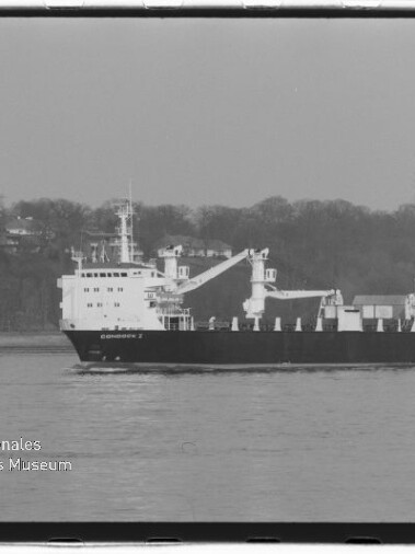 Schwarz-weiß-Foto eines Frachtschiffs mit zwei Ladekränen, das auf ruhigem Wasser vor einem bewaldeten Ufer fährt; links unten Logo und Schriftzug des Internationalen Maritimen Museums Hamburg.