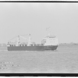 Schwarzweiß-Foto eines Frachtschiffs mit Aufbauten und zwei Ladekränen, das auf ruhigem Meer vor einer flachen Küstenlinie fährt; Wasserzeichen des Internationalen Maritimen Museums Hamburg unten links.