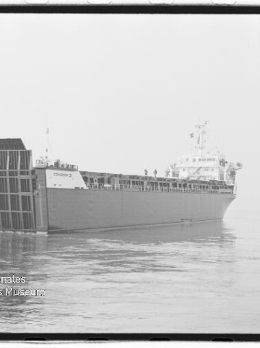 Großes Frachtschiff in Seitenansicht mit abgesenkter Heckrampe, das ruhig auf glatter See liegt; links unten sichtbar das Wasserzeichen des Internationalen Maritimen Museums Hamburg.