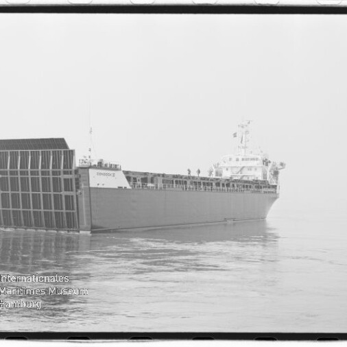 Großes Frachtschiff in Seitenansicht mit abgesenkter Heckrampe, das ruhig auf glatter See liegt; links unten sichtbar das Wasserzeichen des Internationalen Maritimen Museums Hamburg.