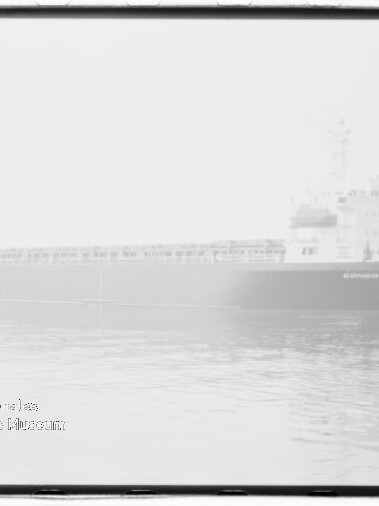 Schwarz-weiß-Foto eines großen Frachtschiffs, das in dichtem Nebel auf ruhigem Wasser treibt, mit leichter Spiegelung und sichtbarem Museumslogo unten links.