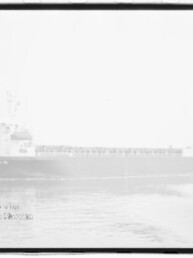 Schwarzweißaufnahme eines seitlich sichtbaren Frachtschiffs auf ruhiger, leicht spiegelnden See in sehr heller/überbelichteter Darstellung, unten links sichtbarer Stempel des Internationalen Maritimen Museums Hamburg