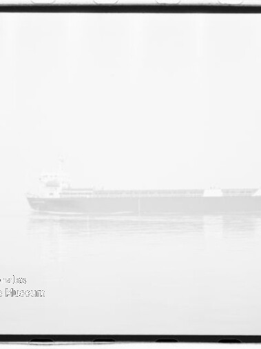 Schwarzweiß-Hochkontrastfoto eines Fracht-/Massengutfrachters, schwach sichtbar und fast überbelichtet, ruhig auf spiegelglattem Meer mit leichter Spiegelung, filmischer Rahmen und Stempel/Wasserzeichen unten links.