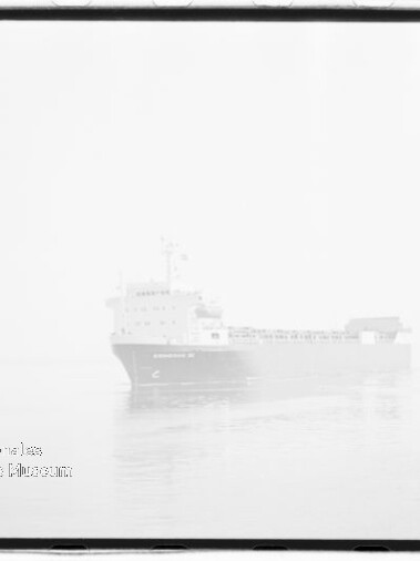 Helles, im Nebel nahezu verschwommenes Frachtschiff auf ruhiger See mit Spiegelung im Wasser und einem Museumsstempel unten links.