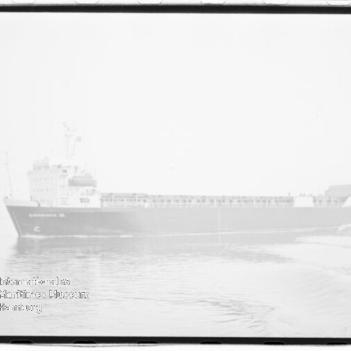 Schwarzweiß-überbelichtetes Foto einer Seitenansicht eines Frachtschiffs auf ruhigem Meer mit leichter Bugwelle, sichtbarem Fotorahmenrand und rundem Wasserzeichen des Internationalen Maritimen Museums Hamburg unten links.