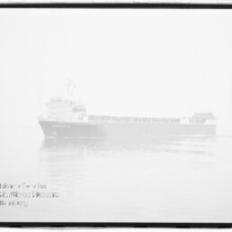 Schwarzweiß-Aufnahme eines großen Frachtschiffs in Seitenansicht auf ruhiger See vor hellem, überbelichtetem Himmel; schwaches Wasserzeichen des Internationalen Maritimen Museums Hamburg unten links und sichtbarer Filmrand.
